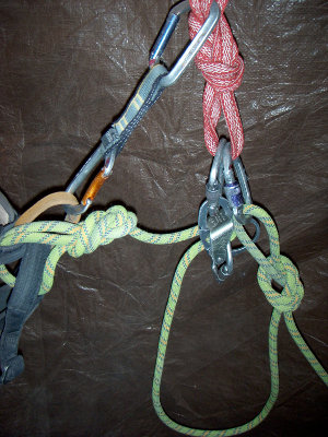 The second climber is secured with an overhand knot behind the belay device and clipped into the anchor.